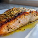 Close-up of a flaky, seasoned air fryer salmon fillet resting in its juices on a white plate.