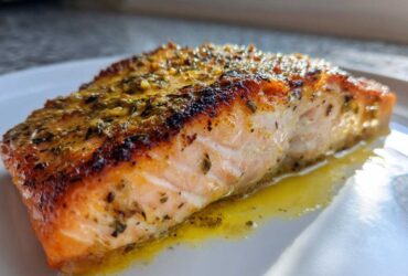 Close-up of a flaky, seasoned air fryer salmon fillet resting in its juices on a white plate.