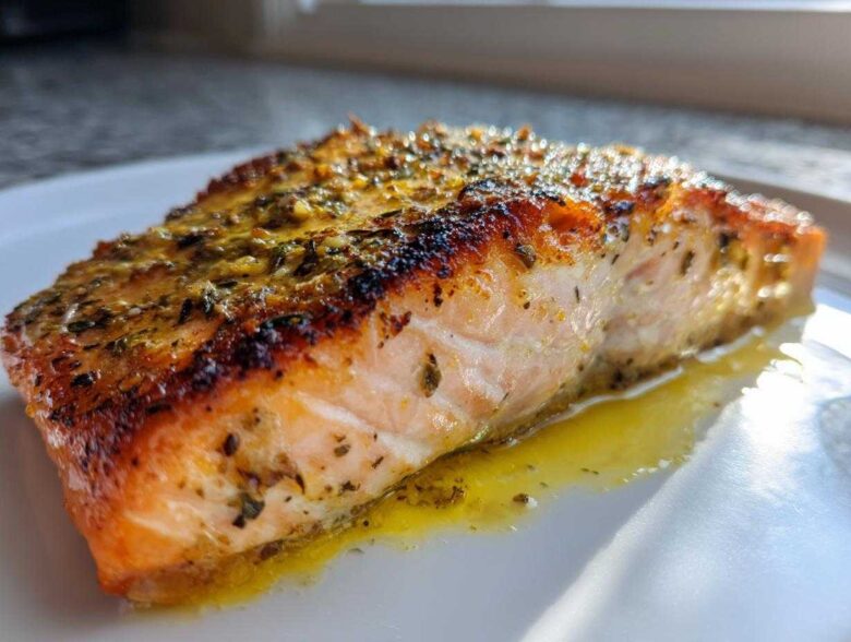 Close-up of a flaky, seasoned air fryer salmon fillet resting in its juices on a white plate.