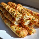 Close-up of golden, cheesy, herb-topped breadsticks stacked on a white plate, showing a fluffy interior.