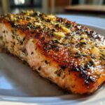 Close-up of a juicy, glazed air fryer salmon fillet topped with garlic and herbs on a white plate.