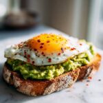 Close-up of perfectly prepared avocado toast topped with a sunny-side-up egg and red pepper flakes.