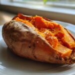 A close-up of a fluffy baked sweet potatoes, split open and sprinkled with coarse sea salt, resting on a white plate.
