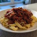 A close-up of rich, meaty bolognese sauce generously spooned over a bed of fresh tagliatelle pasta on a white plate.