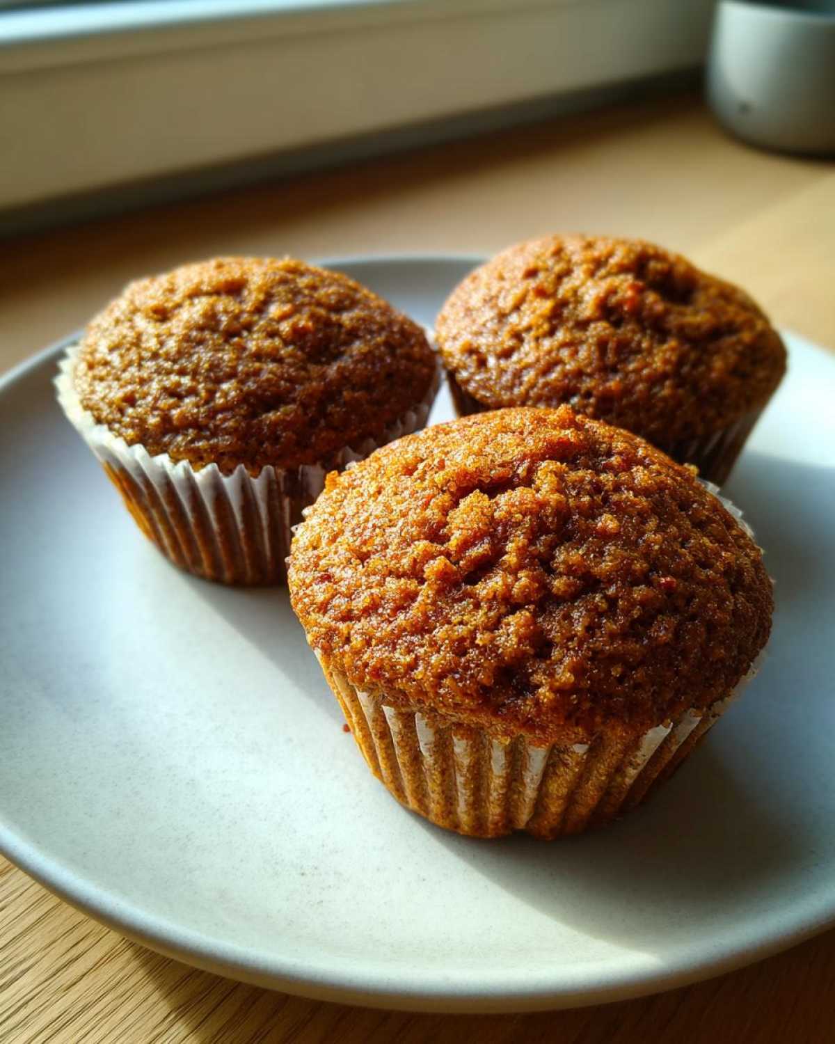 Three golden-brown bran muffins resting in white paper liners on a light grey plate, illuminated by sunlight.