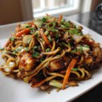 Close-up of a plate piled high with glossy chicken chow mein recipe noodles, carrots, cabbage, and green onions.