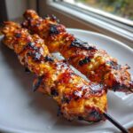 Close-up of two perfectly grilled, glazed chicken skewers with char marks on a white plate.