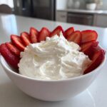 A white bowl filled with creamy fruit dip surrounded by sliced fresh strawberries.