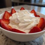 A white bowl filled with creamy fruit dip topped with whipped cream and surrounded by fresh, sliced strawberries.