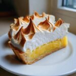 A close-up of a perfect slice of lemon meringue pie showing bright yellow filling and tall, toasted meringue peaks.