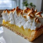 A close-up of a perfect slice of lemon meringue pie showing the crust, bright yellow filling, and tall, torched meringue peaks.