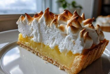 A close-up of a perfect slice of lemon meringue pie showing the crust, bright yellow filling, and tall, torched meringue peaks.