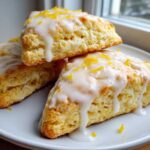 Three triangular lemon scones stacked on a white plate, topped with white glaze and bright yellow lemon zest.