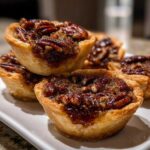 A close-up shot of several gooey, caramelized mini pecan pies stacked and arranged on a white serving plate.