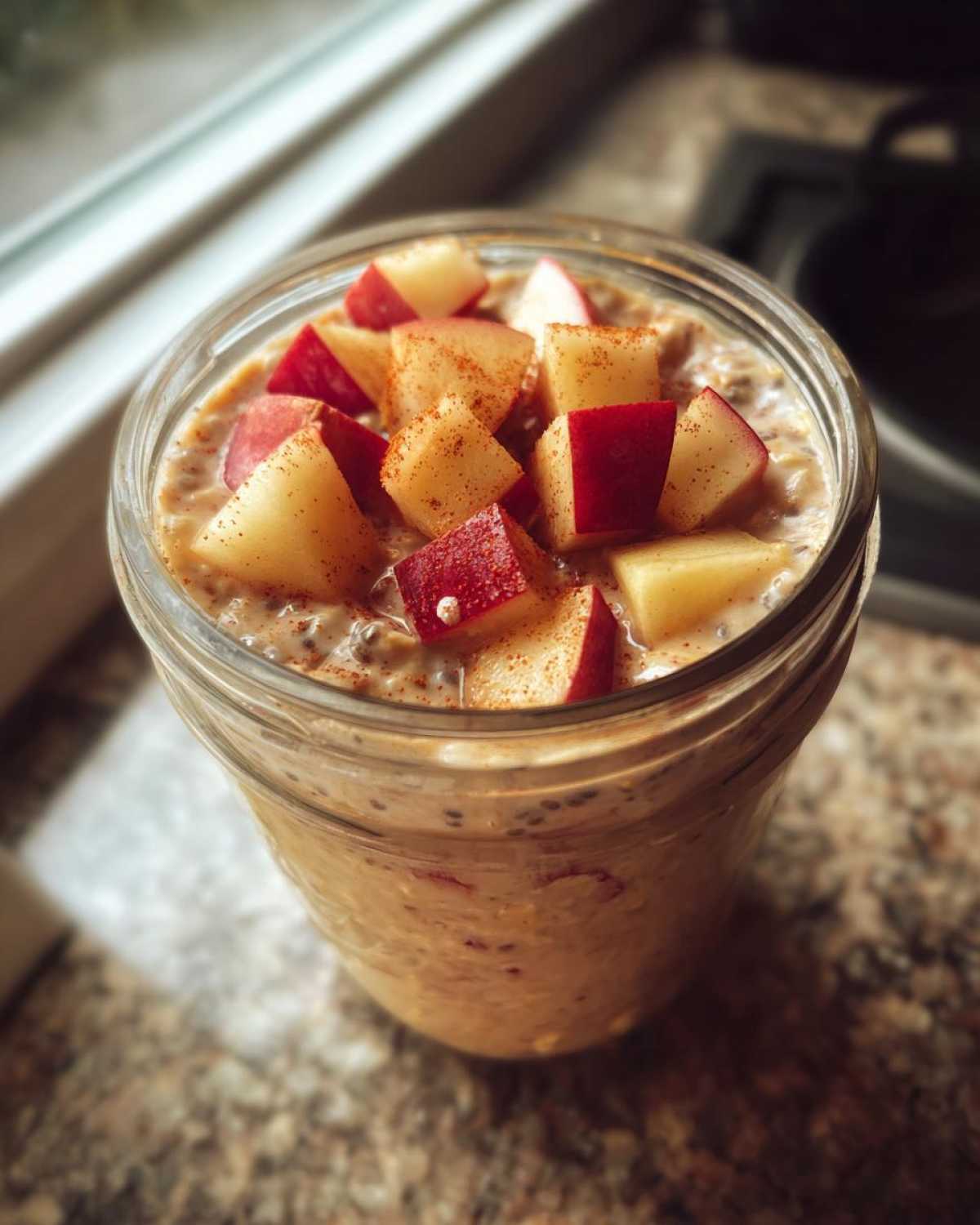 Close-up of apple cinnamon overnight oats in a glass jar, topped with diced red apples and cinnamon.
