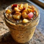 A close-up of apple cinnamon overnight oats topped with diced red apples and cinnamon spice in a glass jar.