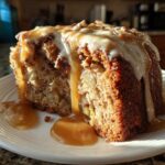 A close-up of a moist slice of apple dapple cake topped with white icing and dripping caramel sauce.