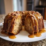 A slice cut from a bundt-style apple dapple cake, heavily drizzled with thick caramel sauce and topped with pecans.