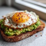 Close-up of delicious avocado toast topped with a sunny-side-up egg and red chili flakes.