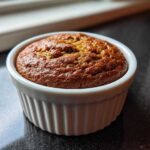 Close-up of a golden brown banana protein muffin baked in a white, fluted ramekin.