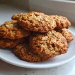 A close-up stack of freshly baked, golden brown oatmeal cookie resting on a small white plate.