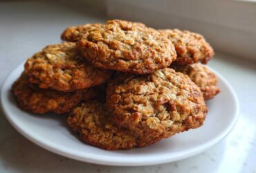 A close-up stack of freshly baked, golden brown oatmeal cookie resting on a small white plate.