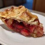 A close-up of a juicy slice of strawberry rhubarb pie showing the bright red filling and golden, flaky top crust.