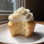 Close-up of a vanilla cupcake with a bite taken out, showing the moist crumb and tall swirl of white vanilla frosting.