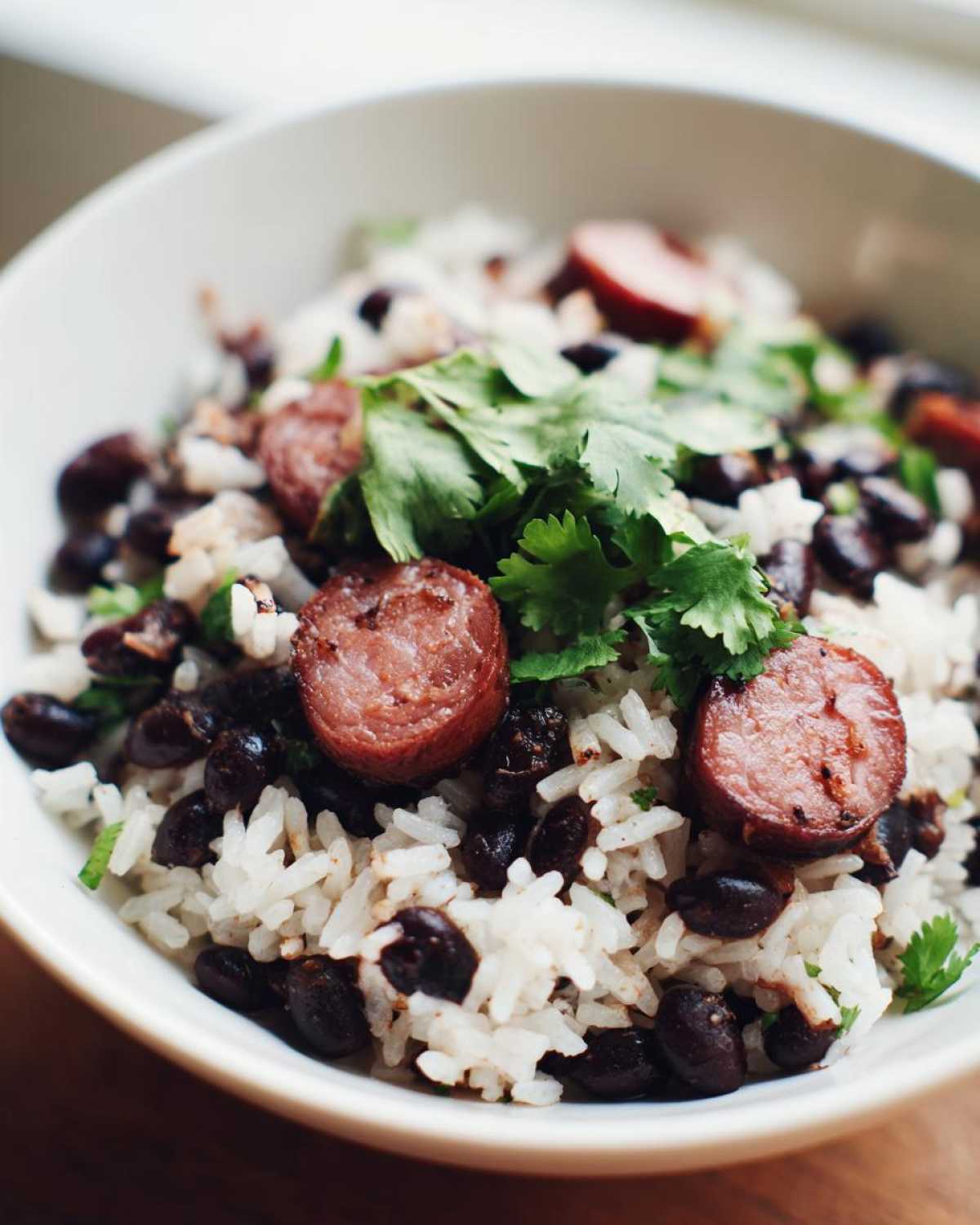 Close-up of a white bowl filled with flavorful black beans and rice, topped with sliced sausage and fresh cilantro.