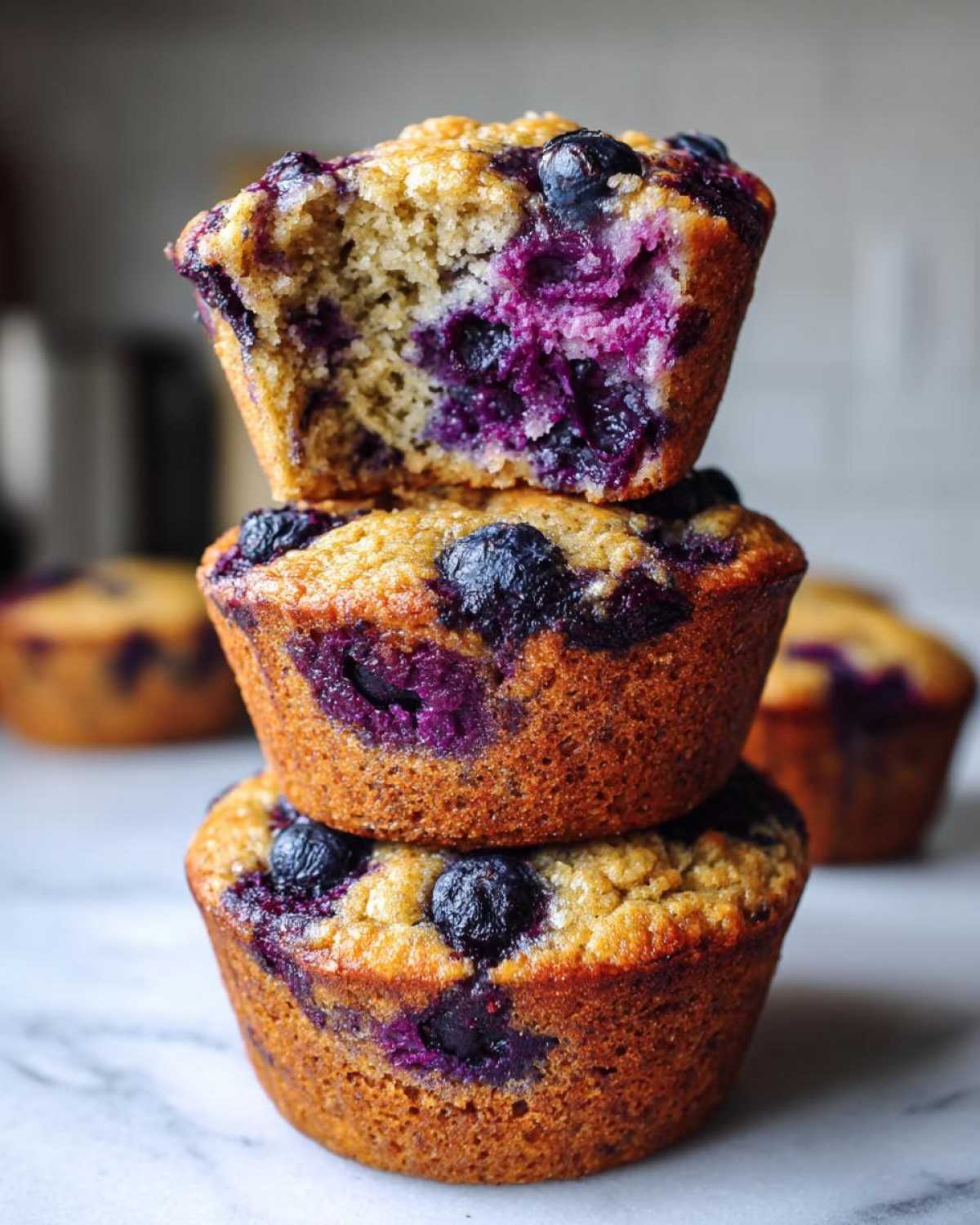 A stack of three golden-brown blueberry protein muffins, with the top one having a bite taken out to show the moist interior.