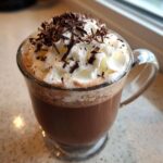 A clear mug filled with rich boozy hot chocolate, topped generously with whipped cream and chocolate shavings.