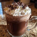 A clear glass mug filled with rich boozy hot chocolate, topped with whipped cream and chocolate shavings.
