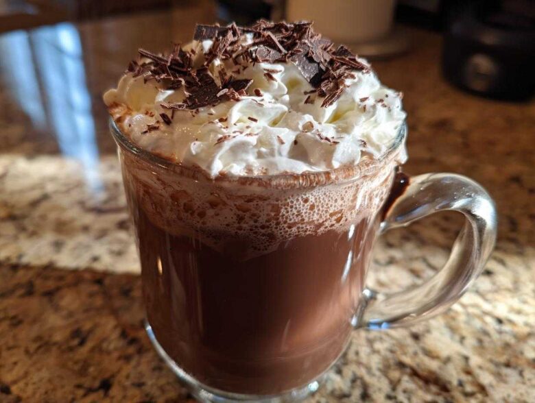 A clear glass mug filled with rich boozy hot chocolate, topped with whipped cream and chocolate shavings.