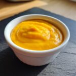 Close-up of thick, bright orange nacho cheese sauce served in a small white dipping bowl on a dark slate surface.