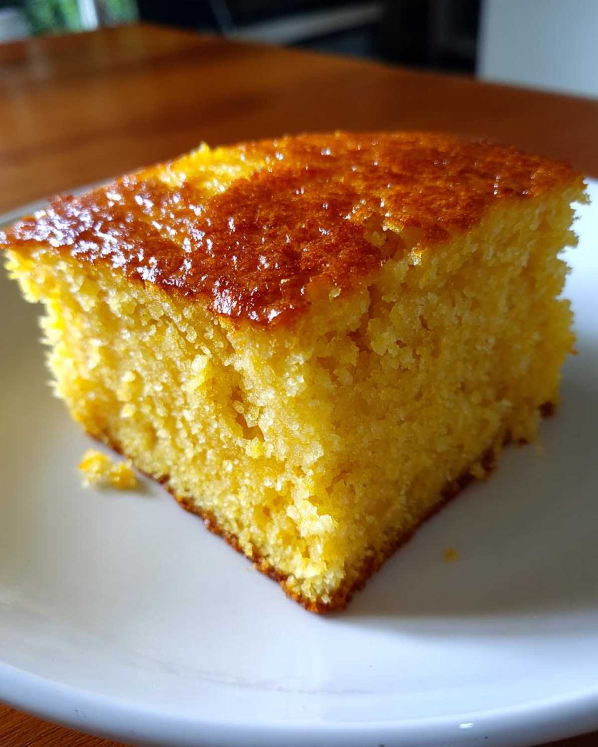 Close-up of a square slice of bright yellow buttermilk cornbread with a shiny, golden-brown top, served on a white plate.