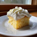 A square slice of yellow cake topped with a generous swirl of light vanilla buttercream frosting on a white plate.