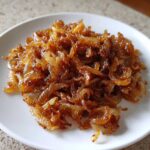 A close-up of a mound of richly browned and glossy caramelized onions served on a white plate.