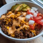 A close-up of a white bowl filled with a cheeseburger bowl featuring seasoned ground beef, melted cheddar, special sauce, pickles, tomatoes, and onions.