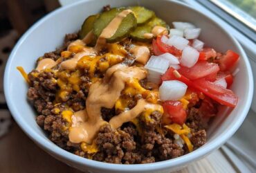 A close-up of a white bowl filled with a cheeseburger bowl featuring seasoned ground beef, melted cheddar, special sauce, pickles, tomatoes, and onions.