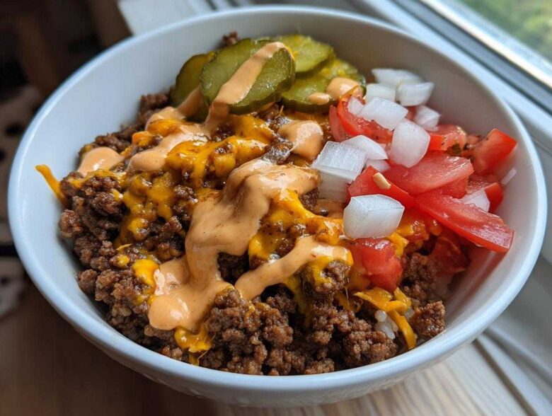 A close-up of a white bowl filled with a cheeseburger bowl featuring seasoned ground beef, melted cheddar, special sauce, pickles, tomatoes, and onions.