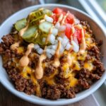 A close-up of a cheeseburger bowl featuring seasoned ground beef, melted cheddar cheese, pickles, onions, tomatoes, and a drizzle of sauce.