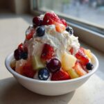 A white bowl overflowing with a creamy cheesecake fruit salad topped generously with fresh strawberries, blueberries, and cherries.