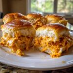 Close-up of cheesy buffalo chicken sliders, one cut in half showing gooey orange filling dripping out.