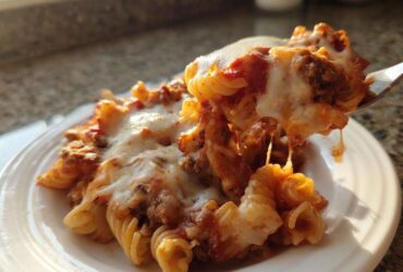 A fork lifts a cheesy, saucy portion of pasta from a plate of pizza casserole, showing melted mozzarella strings.