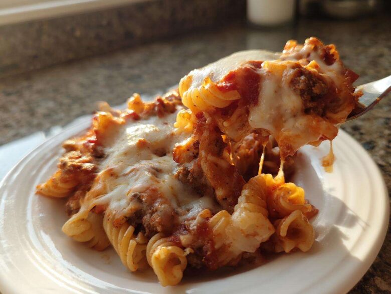 A fork lifts a cheesy, saucy portion of pasta from a plate of pizza casserole, showing melted mozzarella strings.