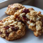 Close-up of chewy cranberry oatmeal cookies, one broken open showing oats, dried cranberries, and white chocolate chips.