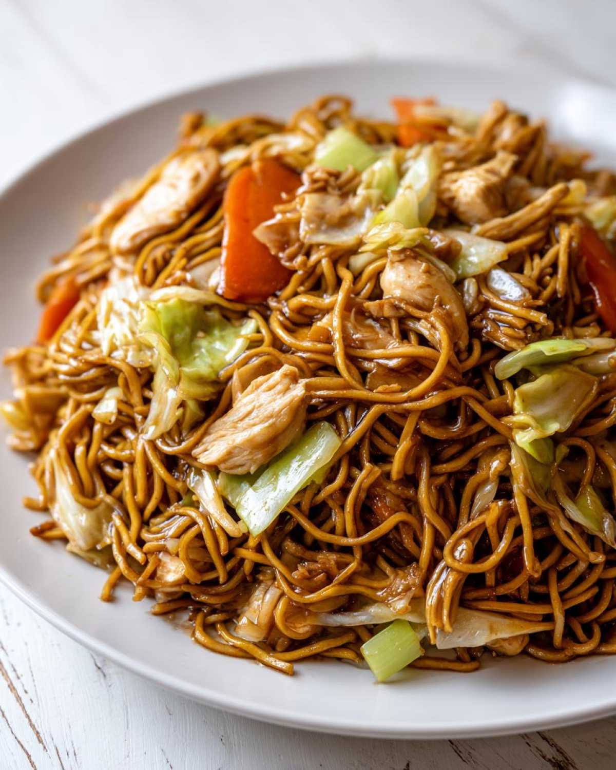 A close-up of a plate piled high with savory chicken chow mein recipe noodles, chicken pieces, cabbage, and carrots.