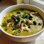 Close-up of a white bowl filled with creamy, green chicken poblano soup recipe, topped with shredded chicken, black beans, and cilantro.