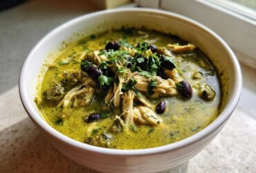 Close-up of a white bowl filled with creamy, green chicken poblano soup recipe, topped with shredded chicken, black beans, and cilantro.