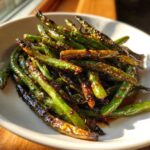 A close-up of blistered, glossy chinese green beans with garlic sauce served in a white bowl.